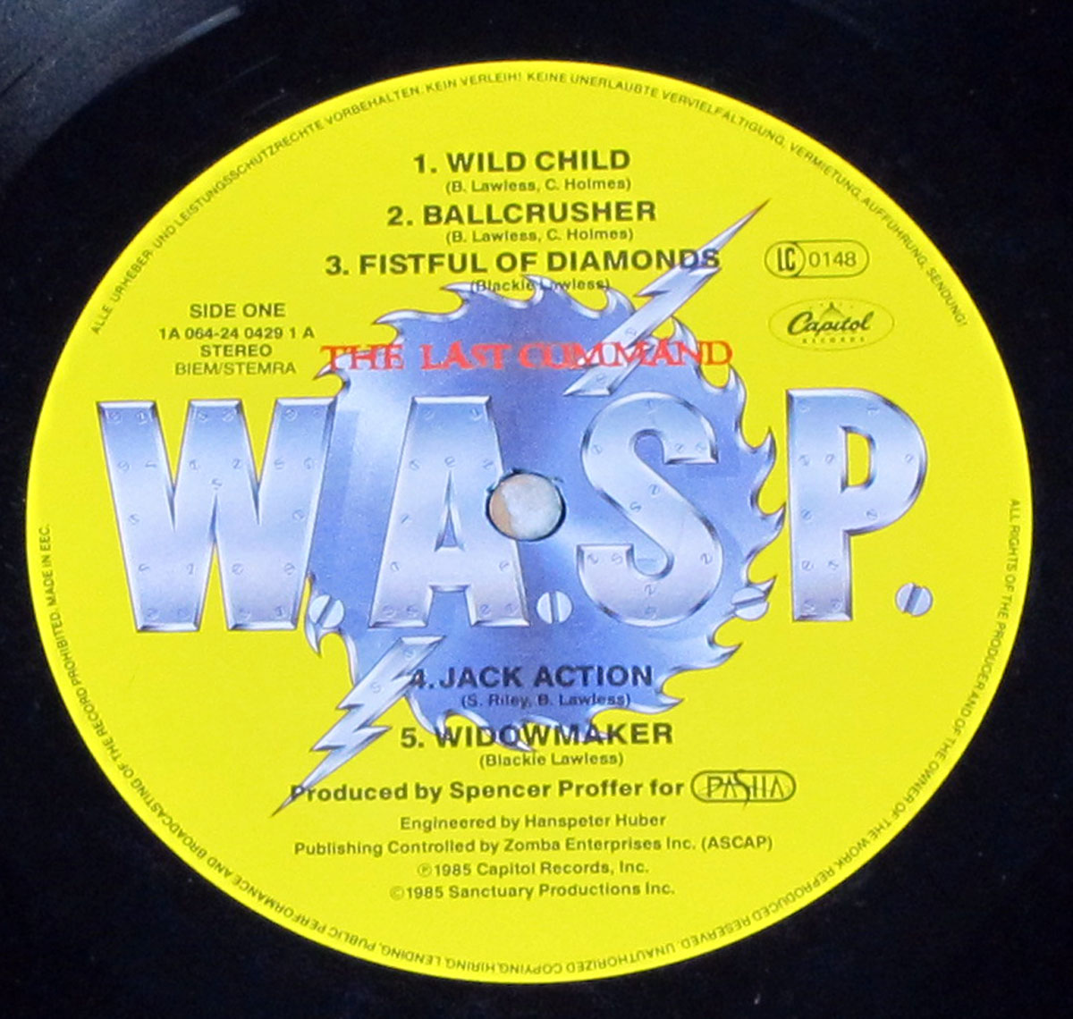 Close up of Side One label for W.A.S.P. "The Last Command": a screaming-bright yellow Capitol label with a huge metallic, riveted W.A.S.P. logo sitting on a jagged blue sawblade shape pierced by lightning bolts, the album title printed in red across the top of the logo, Side One and stereo/BIEM-STEMRA text at left, LC 0148 and the Capitol oval logo at right, plus a numbered track list for Wild Child, Ballcrusher, Fistful of Diamonds, Jack Action, and Widowmaker.